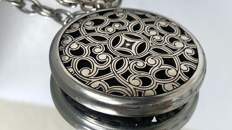 May include: A silver-toned, round pendant with an intricate, swirling, openwork design. The pendant has a black background and is attached to a silver chain. The design is symmetrical and detailed.