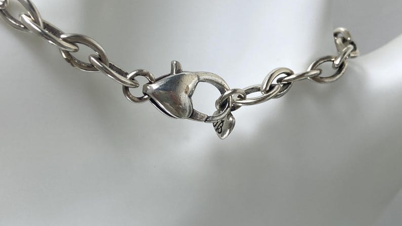 May include: Close-up of a silver necklace clasp and chain. The chain links are oval-shaped and connected to a heart-shaped clasp. The clasp is closed and attached to the chain. The necklace is on a white background.