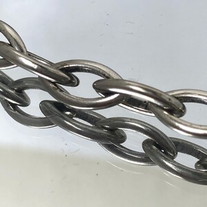 May include: Close-up of a silver-colored chain with oval links. The chain is made of metal and has a shiny, reflective surface. The links are interlocked, creating a strong and durable structure. The chain is likely used for jewelry or other decorative purposes.