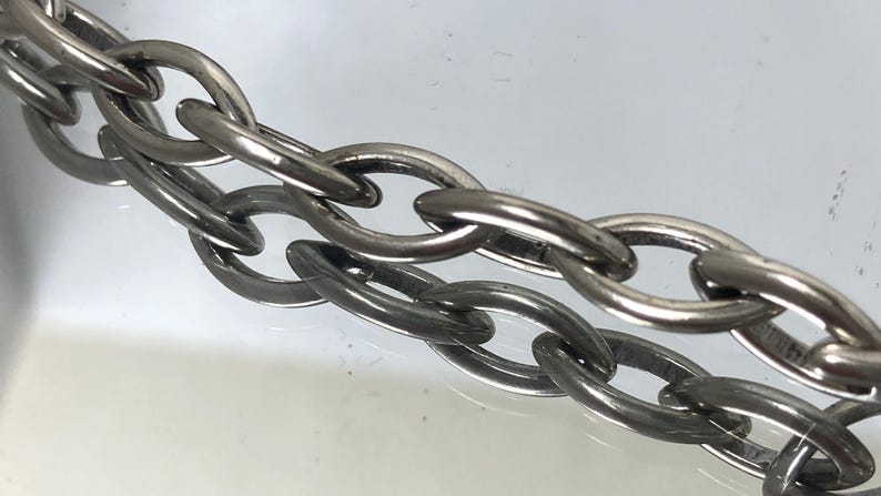 May include: Close-up of a silver-colored chain with oval links. The chain is made of metal and has a shiny, reflective surface. The links are interlocked, creating a strong and durable structure. The chain is likely used for jewelry or other decorative purposes.