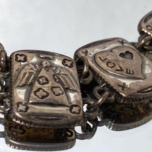 Puede incluir: Una pulsera de dijes de metal plateado con eslabones rectangulares y dise&ntilde;os en relieve. Los eslabones muestran varios s&iacute;mbolos, incluyendo una figura de &aacute;ngel, un coraz&oacute;n con la palabra LOVE y una herradura, unidos por anillos met&aacute;licos.