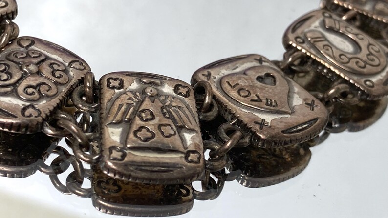 Puede incluir: Una pulsera de dijes de metal plateado con eslabones rectangulares y dise&ntilde;os en relieve. Los eslabones muestran varios s&iacute;mbolos, incluyendo una figura de &aacute;ngel, un coraz&oacute;n con la palabra LOVE y una herradura, unidos por anillos met&aacute;licos.
