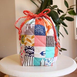 May include: A patchwork drawstring bag with a red and white patterned drawstring closure. The bag is made of various fabric squares in a variety of colours and patterns, including geometric shapes, floral designs, and stripes. The bag is on a white table.