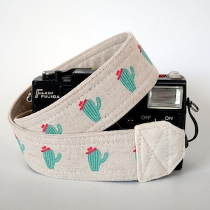 May include: A beige camera strap featuring a repeating pattern of turquoise cacti wearing red hats. The strap is wrapped around a black camera. The camera strap is made of a durable fabric.