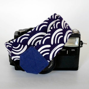 Peut inclure: Une sangle d'appareil photo à motifs bleu marine et blanc est drapée sur un appareil photo noir. La sangle présente un motif de vagues répétées. Un patch bleu marine uni est cousu sur la sangle. L'appareil photo a un flash et un objectif.