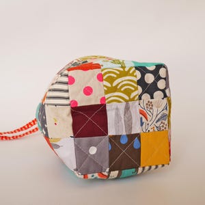 May include: A patchwork cube featuring a mix of patterned fabrics in various colours, such as polka dots, stripes, and floral prints. It has a red and white cord attached. This item is suitable for home decor.