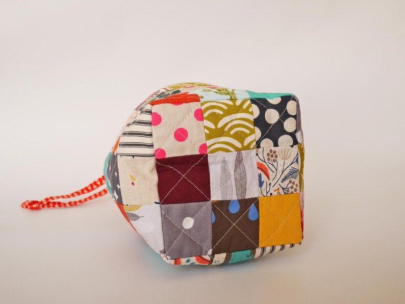 May include: A patchwork cube featuring a mix of patterned fabrics in various colours, such as polka dots, stripes, and floral prints. It has a red and white cord attached. This item is suitable for home decor.