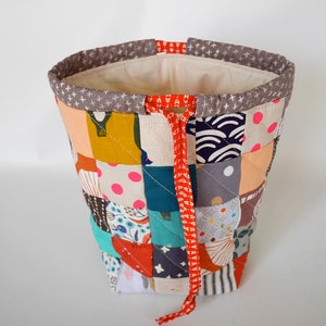 May include: A patchwork fabric bucket bag with a variety of colourful squares. The bag has a grey rim with white cross patterns and a red drawstring. The interior is a light beige fabric.