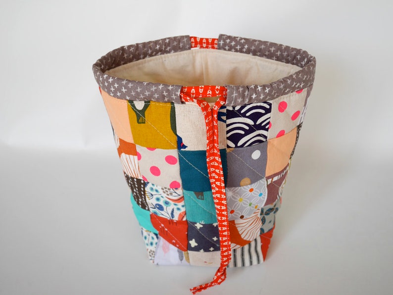May include: A patchwork fabric bucket bag with a variety of colourful squares. The bag has a grey rim with white cross patterns and a red drawstring. The interior is a light beige fabric.