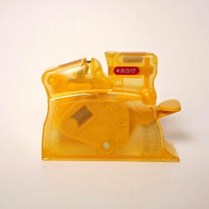 May include: A transparent yellow plastic sewing machine with a red label that has Japanese text. The machine has a lever and a spool holder. The machine is designed for sewing.