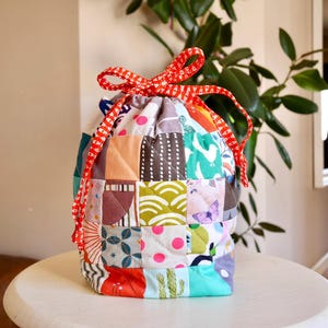 May include: A patchwork drawstring bag with a red and white chequered ribbon closure. The bag is made of various fabric squares in colours like orange, blue, green, and pink. The bag is sitting on a white table.