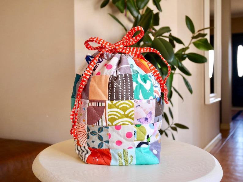 May include: A patchwork drawstring bag with a red and white chequered ribbon closure. The bag is made of various fabric squares in colours like orange, blue, green, and pink. The bag is sitting on a white table.