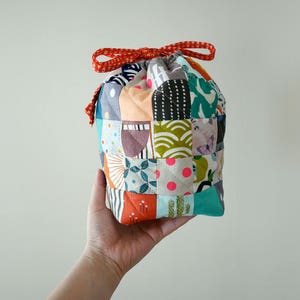 May include: A patchwork drawstring bag with a red ribbon tie. The bag is constructed from a variety of fabric squares in diverse colours and patterns, including floral, geometric, and polka dots. The bag is held in a hand.
