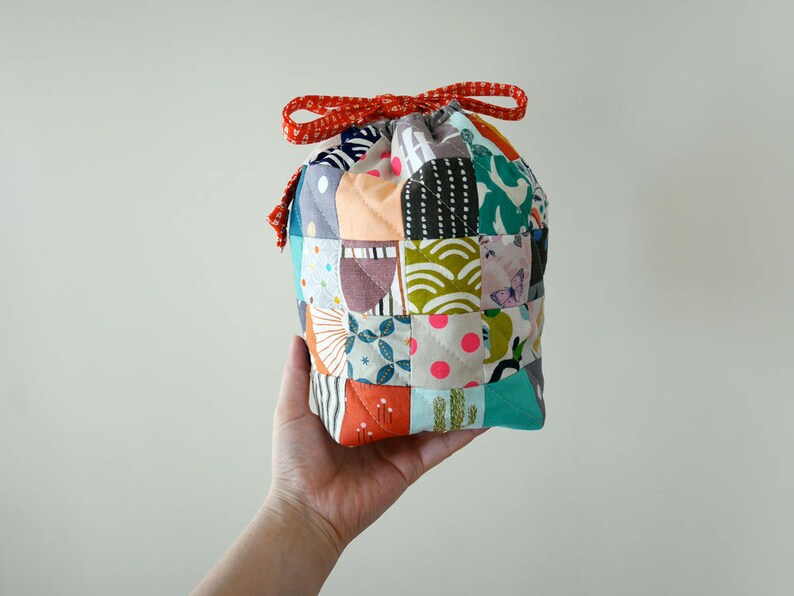 May include: A patchwork drawstring bag with a red ribbon tie. The bag is constructed from a variety of fabric squares in diverse colours and patterns, including floral, geometric, and polka dots. The bag is held in a hand.