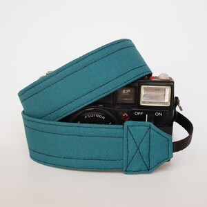 May include: Teal camera strap wrapped around a black vintage camera. The strap is made of a durable fabric with black stitching. The camera has the word "FUJINON" printed on it.