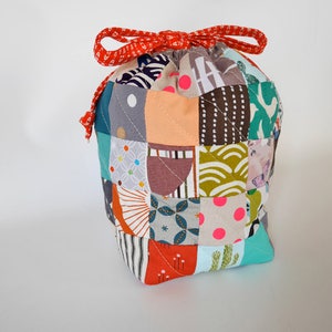 May include: A patchwork drawstring bag featuring a variety of patterned squares in shades of teal, orange, and brown. The bag is secured with a red patterned ribbon. A unique and colourful accessory.