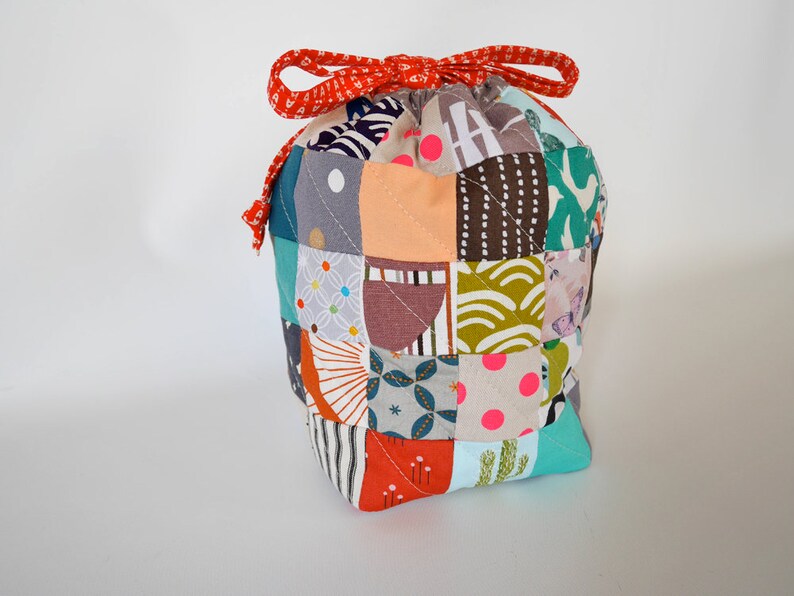 May include: A patchwork drawstring bag featuring a variety of patterned squares in shades of teal, orange, and brown. The bag is secured with a red patterned ribbon. A unique and colourful accessory.