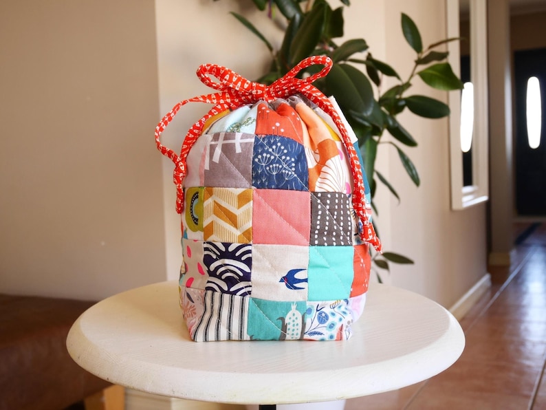 May include: A patchwork drawstring bag with a red and white patterned drawstring closure. The bag is made of various fabric squares in a variety of colours and patterns, including geometric shapes, floral designs, and stripes. The bag is on a white table.