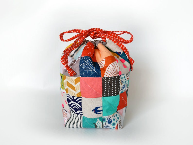 May include: A patchwork drawstring bag with a red patterned drawstring. The bag is made of various fabric squares in colours like teal, pink, orange, and grey. The bag has a rounded shape and is likely used for storage.