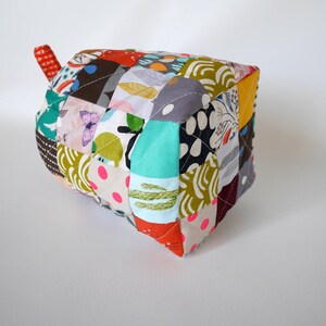 May include: A handmade, patchwork fabric pot holder in a cube shape. The pot holder features a variety of colourful fabric scraps with floral, polka dot, and abstract patterns. A small, red fabric loop is attached to one side.