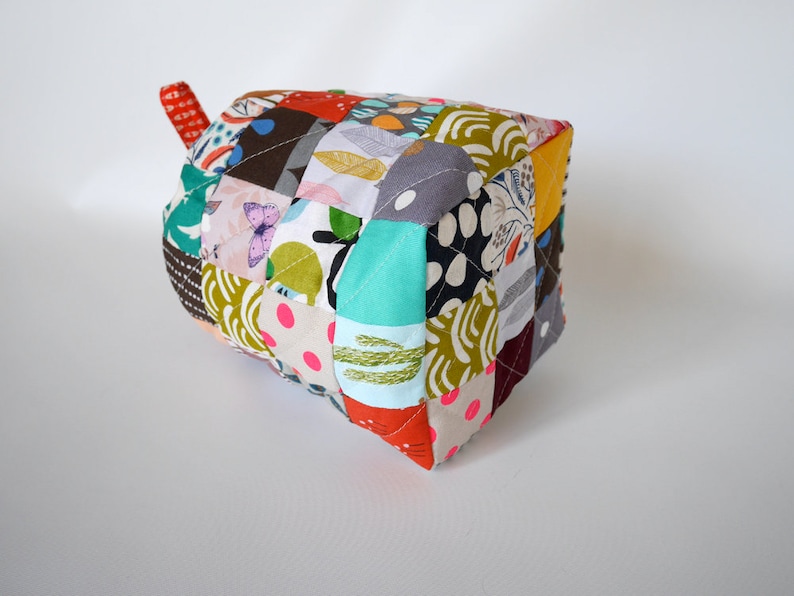 May include: A handmade, patchwork fabric pot holder in a cube shape. The pot holder features a variety of colourful fabric scraps with floral, polka dot, and abstract patterns. A small, red fabric loop is attached to one side.