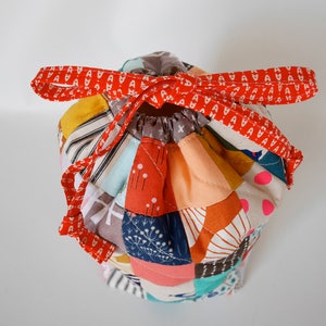 May include: A patchwork drawstring bag in various colours, with a red patterned ribbon tie. The bag is made from a variety of fabrics, including orange, yellow, blue, and pink. The bag is designed to be closed with a drawstring.
