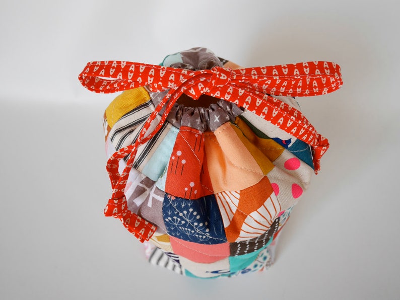 May include: A patchwork drawstring bag in various colours, with a red patterned ribbon tie. The bag is made from a variety of fabrics, including orange, yellow, blue, and pink. The bag is designed to be closed with a drawstring.