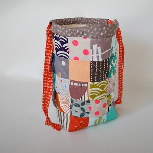 May include: A patchwork fabric drawstring bag with a variety of colourful square patterns. The bag has a grey top with white cross patterns and red straps. The bag is approximately 20 cm tall.