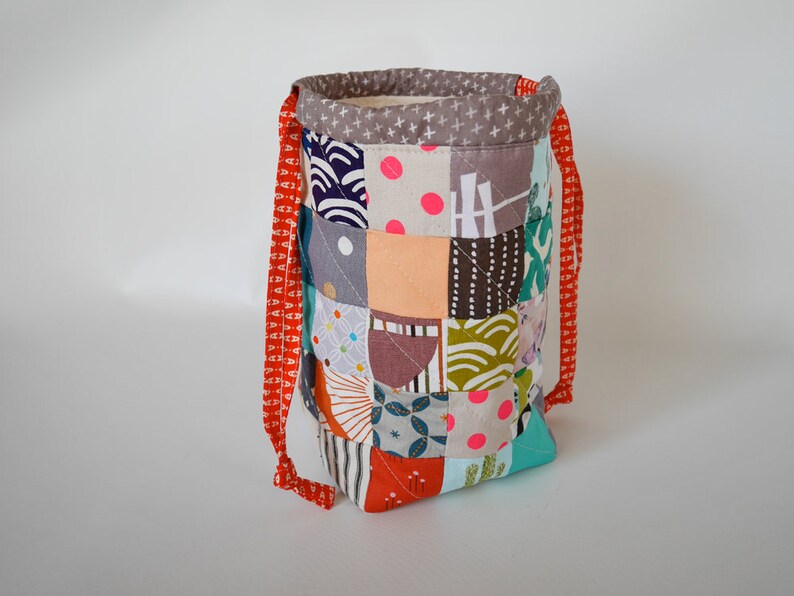 May include: A patchwork fabric drawstring bag with a variety of colourful square patterns. The bag has a grey top with white cross patterns and red straps. The bag is approximately 20 cm tall.