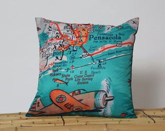 May include: A turquoise and orange throw pillow with a vintage map of Pensacola, Florida. The map shows roads, towns, and points of interest. A red and orange airplane is flying over the map.