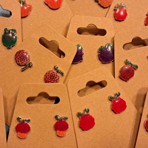May include: A collection of colorful enamel earrings on brown cardboard display cards. The earrings feature various fruit and flower designs, including strawberries, raspberries, and dahlias. The earrings are in shades of red, purple, green, and orange.