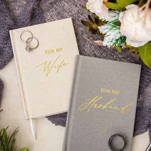 May include: Two journals with linen covers, one beige and one gray. The covers have gold lettering that reads "For My Wife" and "For My Husband".