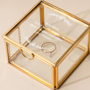 May include: A clear glass jewelry box with gold-colored metal trim. The box contains a ring with a pearl and has the text "Dear Mom. Out of all the mom out there, I'm glad your mine. Love, Pamela" etched on the lid.