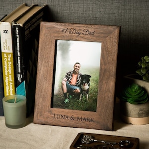 May include: A dark brown wooden picture frame with a photo of a man and a dog. The frame has the text "#1 Dog Dad" and "LUNA & MARK" engraved on it.