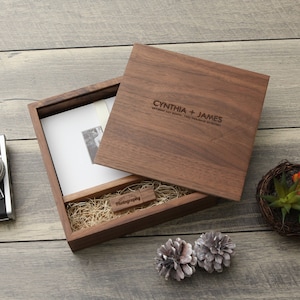 May include: A wooden box with an open lid, revealing a photograph and a USB drive. The box is engraved with the names "CYNTHIA + JAMES" and the date. The box is filled with straw and is on a wooden surface.
