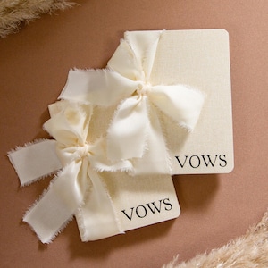 May include: Two ivory-colored cards with the word "VOWS" printed in black, tied with ivory fabric bows. The cards have a textured surface and are arranged on a brown background, with dried ornamental grass in the background.