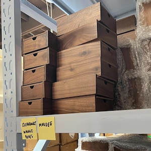 May include: A collection of wooden walnut boxes with drawers, stacked on shelves. The boxes vary in size and are made of a rich, dark wood. The text "CLEARANCE WALNUT BOXES" is visible on a yellow sticky note.