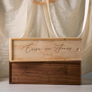 May include: Two wooden boxes stacked on top of each other. The top box is light wood with the text "Crisa and Jonas 12.04.22" engraved on it. The bottom box is dark wood with the text "Jennie and Mike 11.11.22" engraved on it.