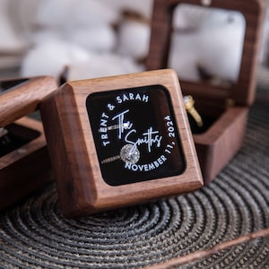 May include: A wooden ring box with a clear glass top. The box is engraved with the text "Trent & Sarah The Smiths November 11, 2024".
