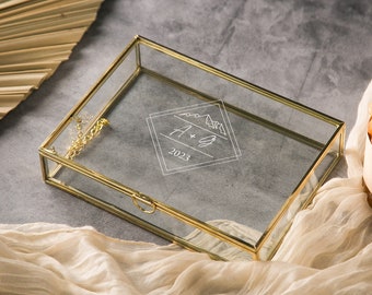 May include: A gold-framed glass box with a clear interior. The box has a hinged lid and a small chain attached to the lid. The box has a white inscription that reads "A + S 2023" inside a diamond shape.