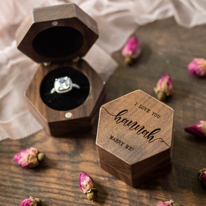 May include: A wooden hexagonal ring box with the words "I love you hannah marry me?" engraved on the lid. The box is open, revealing a diamond ring. Dried pink rosebuds are scattered around the box on a wooden surface.
