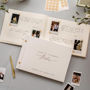 May include: A white wedding guest book with gold accents, open to display handwritten notes and attached polaroid photos. The cover reads "Camille & Andrew Parker" with the date and location below. A gold pen and additional photos are scattered around.
