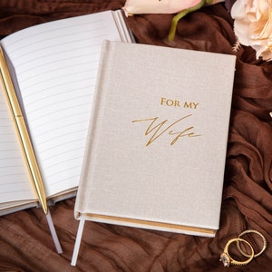 May include: A beige linen journal with gold lettering that reads "For My Wife". The journal is open to a blank page with a gold pen lying on top. The journal is sitting on a brown fabric surface with a few white roses in the background.