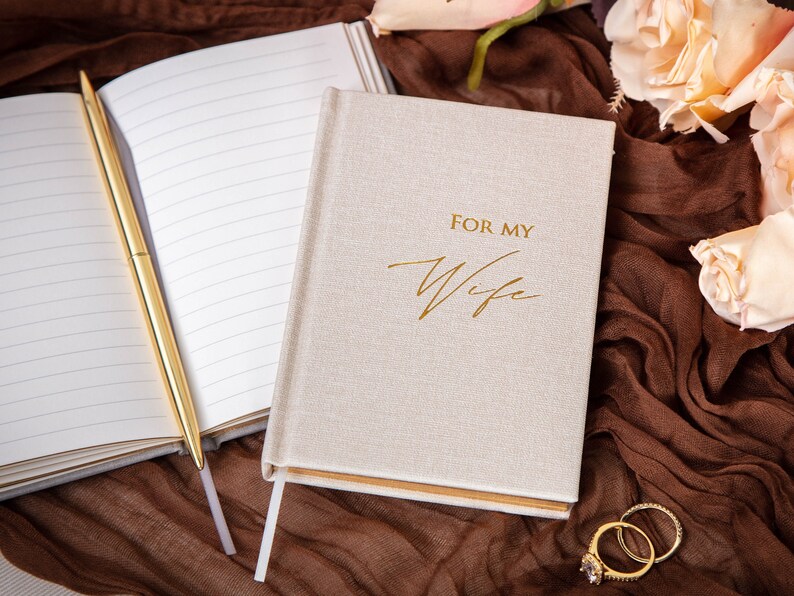 May include: A beige linen journal with gold lettering that reads "For My Wife". The journal is open to a blank page with a gold pen lying on top. The journal is sitting on a brown fabric surface with a few white roses in the background.