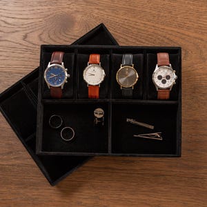 May include: A black velvet watch and accessory organizer with four watches, two rings, and two tie clips. The watches have leather bands in brown and orange, with silver and gold watch faces. The organizer sits on a wooden surface.