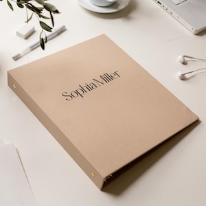 May include: A tan binder with the name "Sophia Miller" printed in black. The binder is on a white surface with a pen, eraser, and earbuds nearby. The binder has gold-colored metal rings.