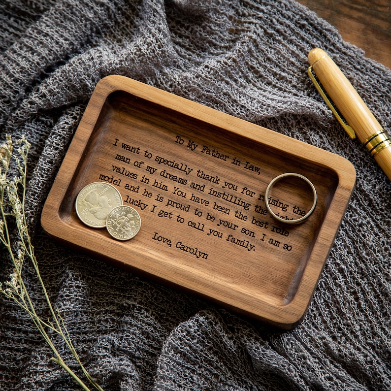 K&ouml;nnte beinhalten: Ein rechteckiges Holztablett mit abgerundeten Ecken und einer Gravur: To My Father in Law, I want to specially thank you for raising the man of my dreams and instilling the right values in him. Love, Carolyn. Enth&auml;lt einen Ring und einige M&uuml;nzen.