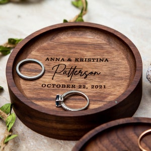 May include: A wooden ring dish with the inscription "Anna & Kristina Patterson October 22, 2021". Two silver rings are resting on the dish.