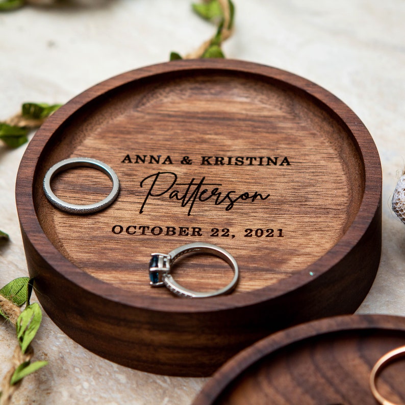 May include: A wooden ring dish with the inscription "Anna & Kristina Patterson October 22, 2021". Two silver rings are resting on the dish.