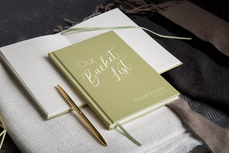 Puede incluir: Un cuaderno verde con las palabras "Our Bucket List" escritas en cursiva blanca. El cuaderno est&aacute; personalizado con los nombres "Russell & Marie" y la fecha "10.02.23".